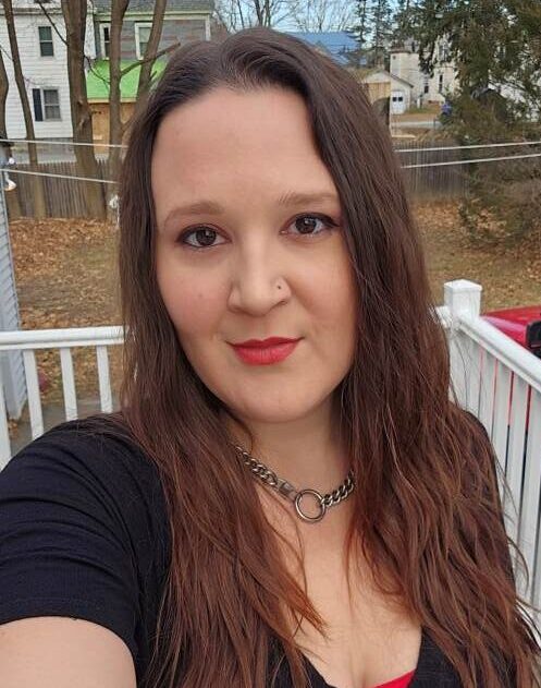 A woman with long wavy brown hair looks into the camera. She wears a chain necklace and a black shirt.